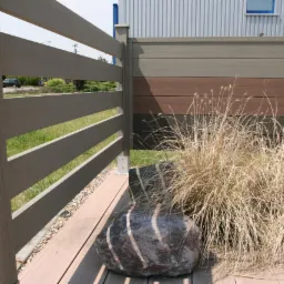 Nowoczesne ogrodzenie kompozytowe z poziomymi panelami w odcieniach szarości i brązu, z elementami dekoracyjnymi w postaci kamienia i trawy ozdobnej na drewnianym tarasie.