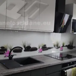 Szklany panel kuchenny z motywem zen: czarne kamienie, trawa i różowe lotosy na białym tle, czarny zlew i płyta indukcyjna w nowoczesnej, szarej kuchni z białymi szafkami.