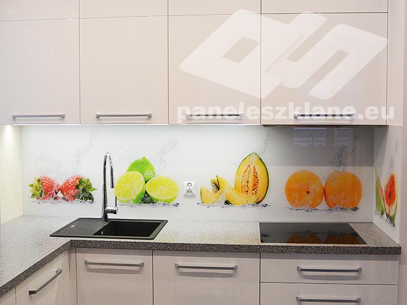 Szklany panel kuchenny z motywem owoców (truskawki, limonki, melon kantalupa, pomarańcze) zamontowany nad blatem z czarnym zlewozmywakiem i płytą indukcyjną; białe szafki wiszące i stojące z szarym...