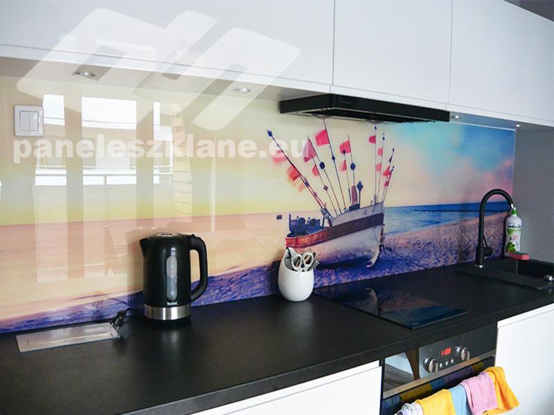Szklany panel kuchenny z motywem łodzi rybackiej na plaży o zachodzie słońca, czarny czajnik elektryczny na blacie, widoczne logo firmy na panelu.