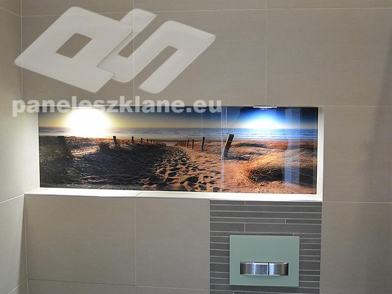 Szklany panel ścienny w łazience z motywem plaży o zachodzie słońca, wkomponowany w ceramiczne płytki, widoczne logo firmy na górze.