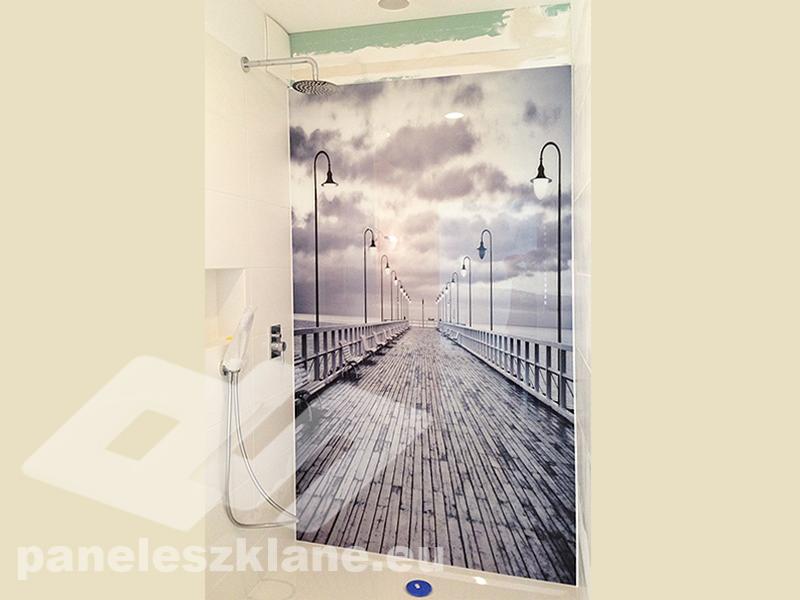 Szklana ściana kabiny prysznicowej z nadrukowanym motywem molo z latarniami i pochmurnym niebem, widoczna bateria prysznicowa i słuchawka.