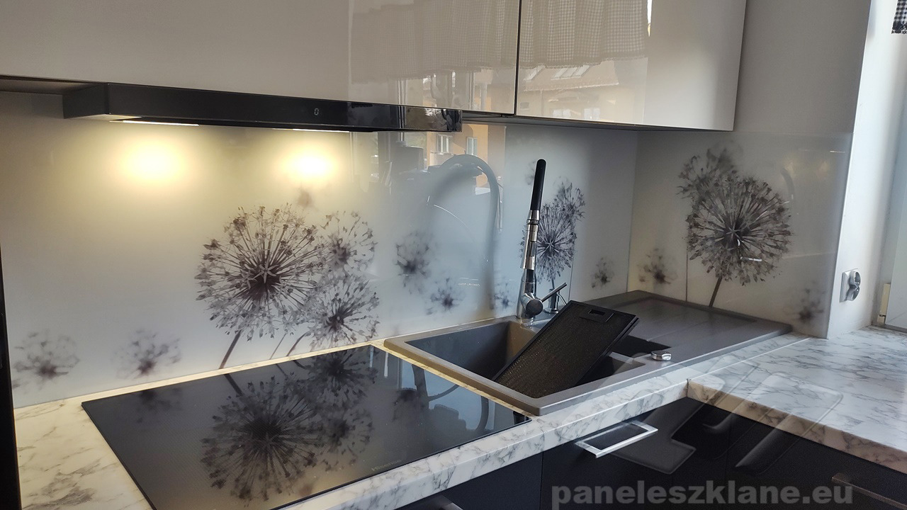 Szklany panel kuchenny z motywem dmuchawców, zamontowany nad płytą indukcyjną i zlewem, oświetlony od dołu, z widocznymi białymi szafkami wiszącymi.