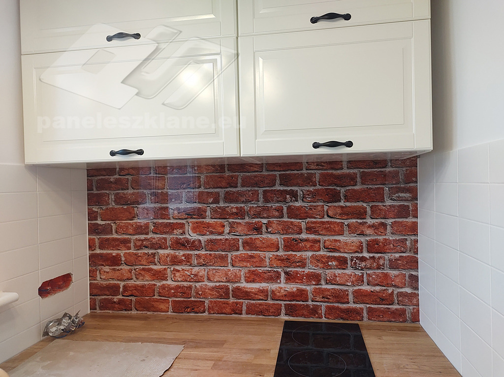 Szklany panel ścienny z motywem czerwonej cegły, zamontowany między blatem kuchennym a wiszącymi, kremowymi szafkami z czarnymi uchwytami. Widoczne białe płytki na ścianie z lewej strony i płyta...