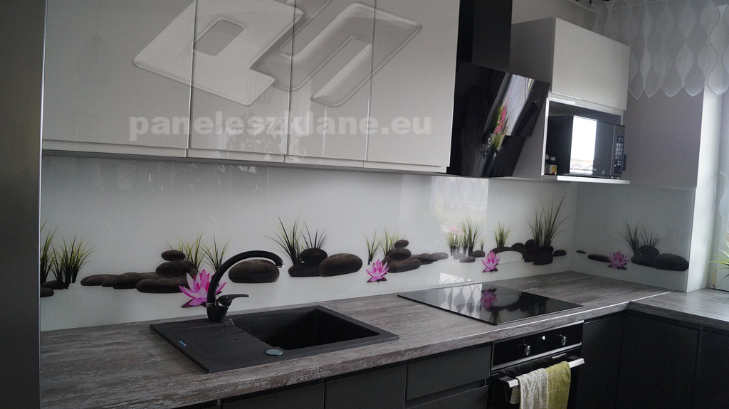 Szklany panel kuchenny z motywem zen: czarne kamienie, trawa i różowe lotosy na białym tle, czarny zlew i płyta indukcyjna w nowoczesnej, szarej kuchni z białymi szafkami.