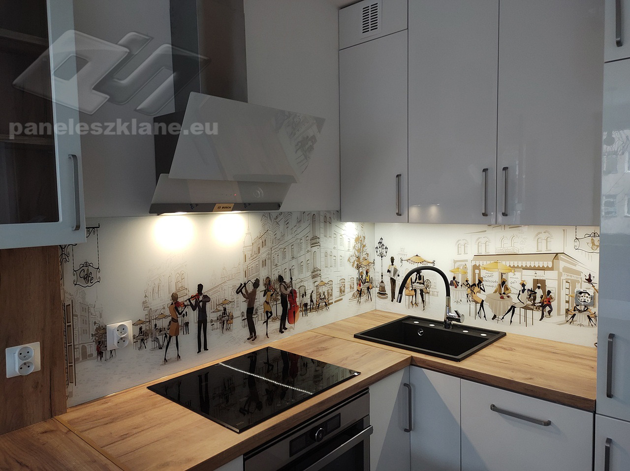 Szklany panel kuchenny z grafiką przedstawiającą uliczną scenę z muzykami i kawiarniami w stylu retro, oświetlony punktowo, z widoczną płytą indukcyjną i zlewozmywakiem.