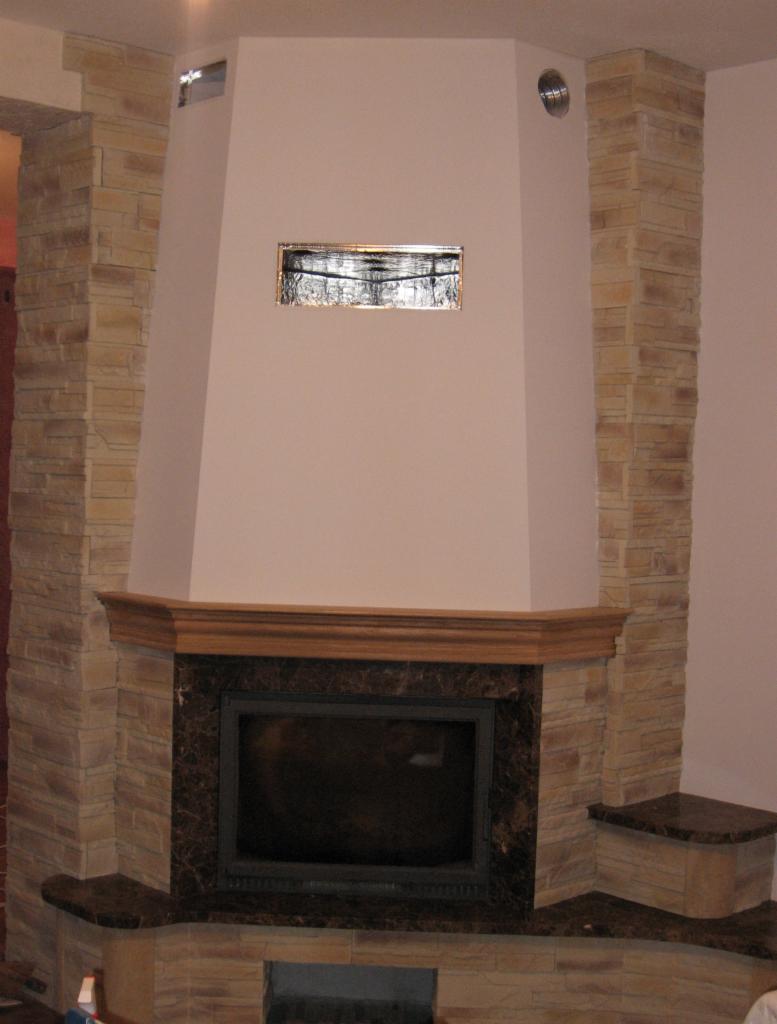 Obudowa kominka wykończona kamieniem dekoracyjnym w odcieniach beżu i brązu. Widoczny wkład kominkowy, drewniany gzyms i fragment ściany z wentylacją.