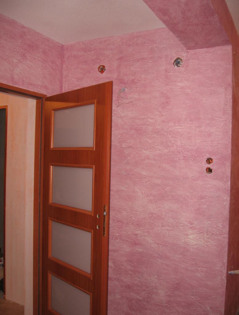 Różowa ściana z dekoracyjną fakturą, częściowo widoczne drewniane drzwi z trzema szybami. Widoczne gniazdka elektryczne bez osprzętu.