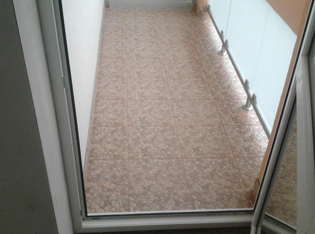 Wykończony balkon z beżową, wzorzystą terakotą ułożoną w kwadraty, widok z góry przez otwarte drzwi balkonowe.