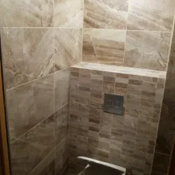 Wykończona toaleta w łazience z beżowymi płytkami imitującymi marmur na ścianach i mozaiką w strefie zabudowy stelaża podtynkowego. Uwagę zwraca nowoczesny przycisk spłukujący.