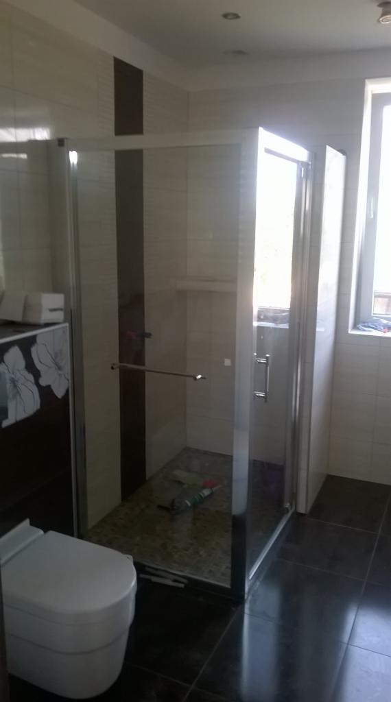 Wykończona łazienka z kabiną prysznicową typu walk-in, ciemną podłogą i jasnymi płytkami z ciemnym pionowym pasem; widoczny biały sedes i narzędzia wewnątrz kabiny.