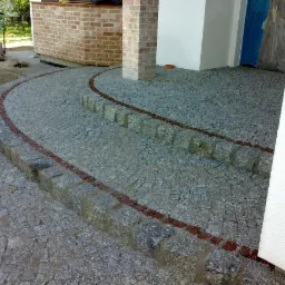 Ułożona granitowa kostka tworząca schody z czerwoną linią oddzielającą stopnie, z ubijarką w tle. Widok na budynek z ceglanym wykończeniem.