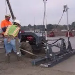 Praca na budowie: robotnicy w kaskach i kamizelkach obsługują maszynę do wylewania posadzki na rozległym placu budowy, z widocznym sprzętem i elementami konstrukcyjnymi.