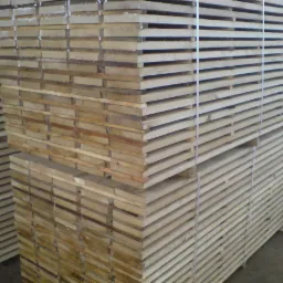 Stosy jasnego drewna budowlanego ułożone w równych rzędach, spięte taśmami. Widoczne detale struktury drewna i przestrzenie między deskami. Tło lekko rozmyte.