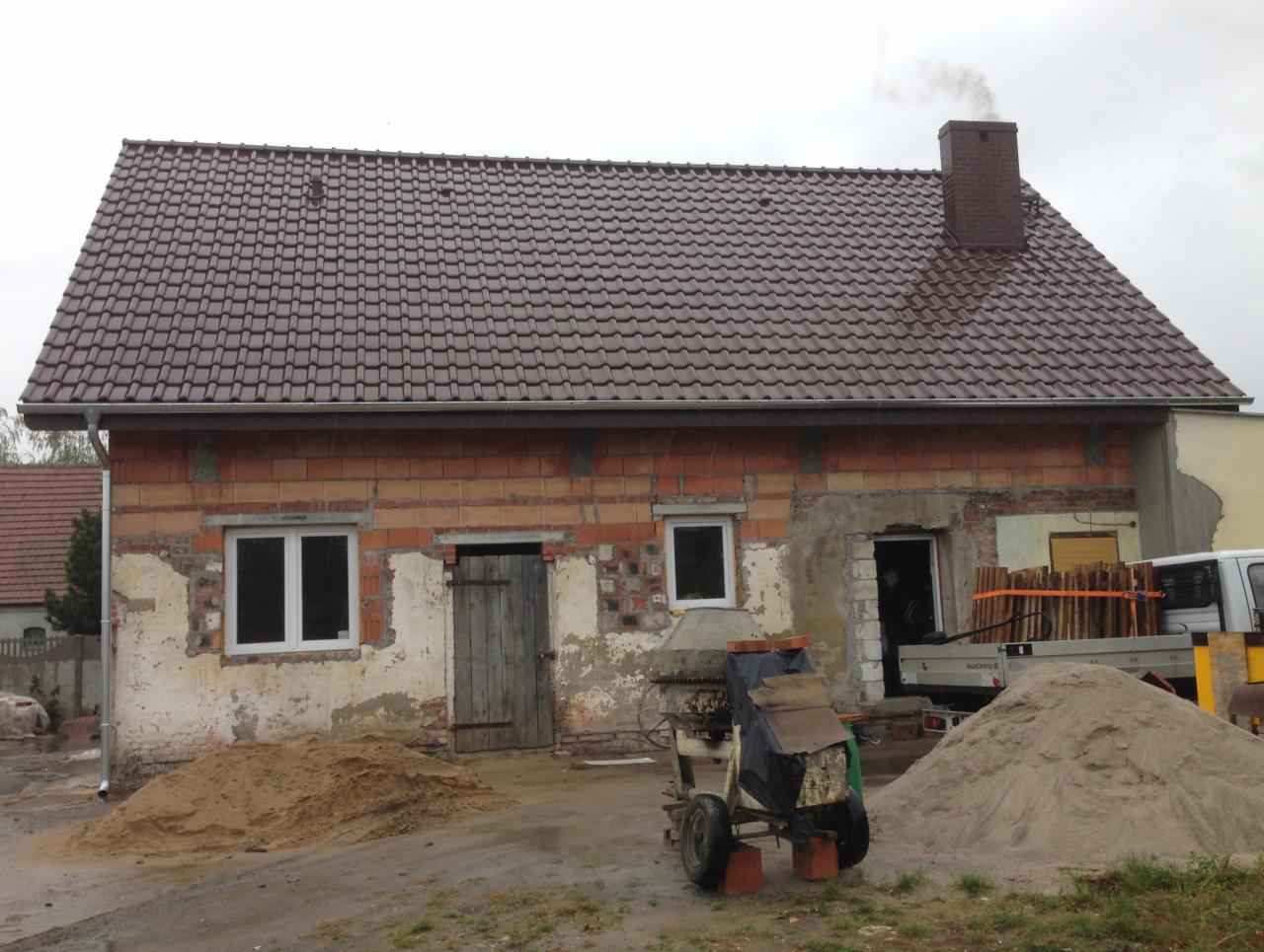 Budynek w trakcie remontu z nowym brązowym dachem, widoczna mieszarka do betonu i sterty piasku, fragment rynny i komin z dymem.