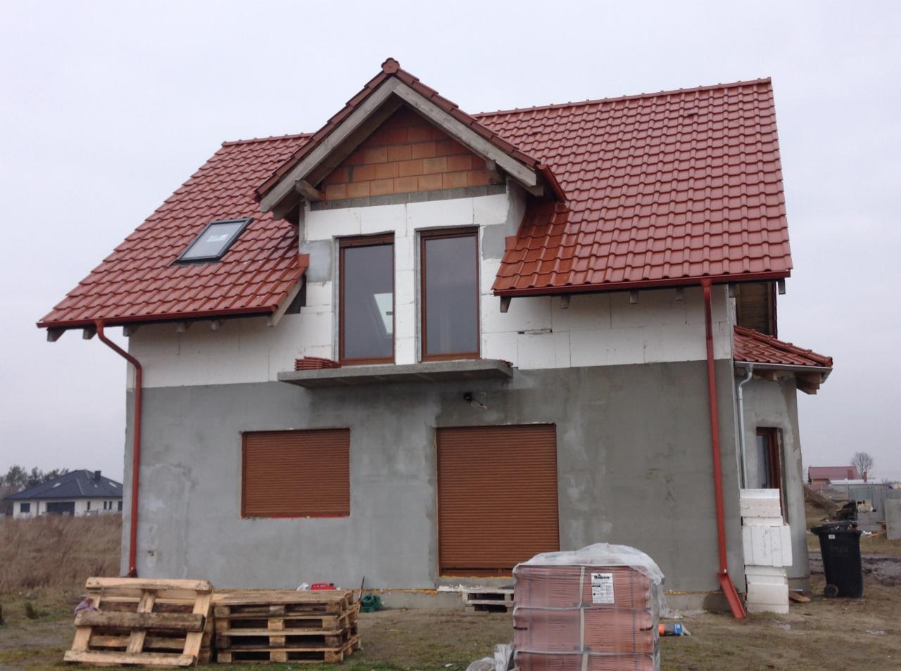 Dom w trakcie budowy z częściowo ukończonym czerwonym dachem z dachówki ceramicznej, widoczne rynny, okna z roletami zewnętrznymi i materiały budowlane na paletach.