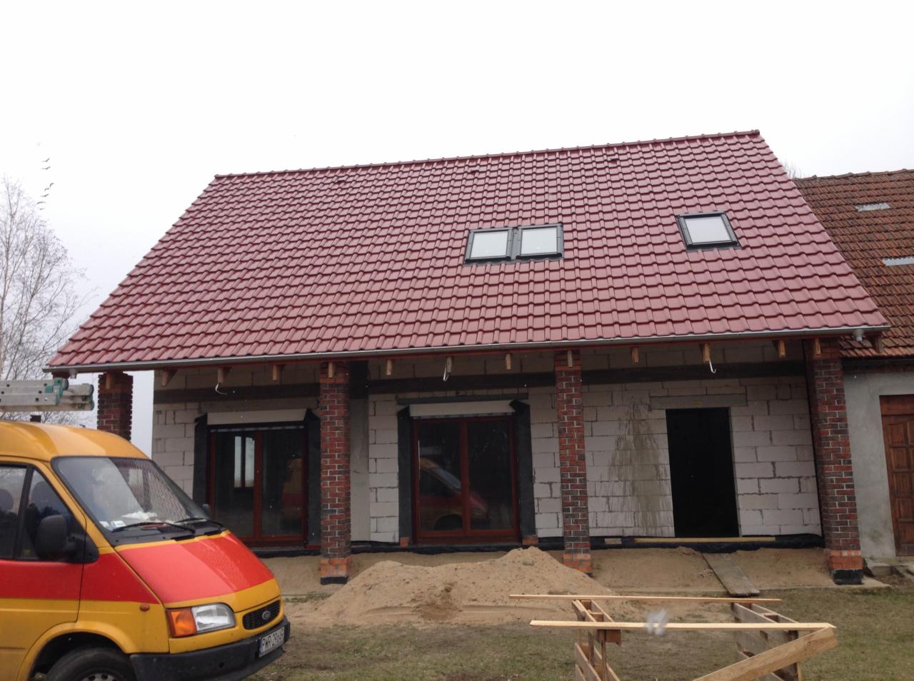 Dom w trakcie budowy z nowym dachem z czerwonej dachówki ceramicznej i oknami dachowymi, widoczny żółto-czerwony samochód dostawczy i częściowo zadaszone wejście.