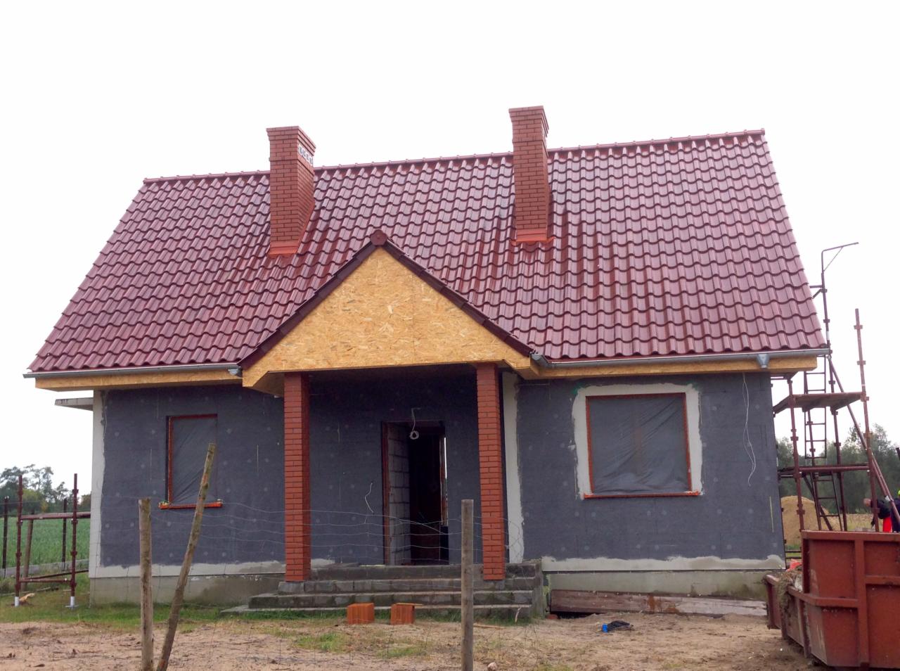 Dom w trakcie budowy z nowym, czerwonym dachem z dachówki ceramicznej, dwoma kominami z cegły, widoczne rusztowania i przygotowane materiały budowlane.