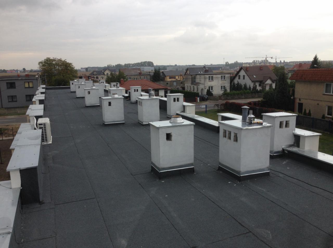 Płaski dach pokryty czarną papą z licznymi, symetrycznie rozmieszczonymi, betonowymi kominami wentylacyjnymi pomalowanymi na biało, widok na zabudowania mieszkalne w tle.