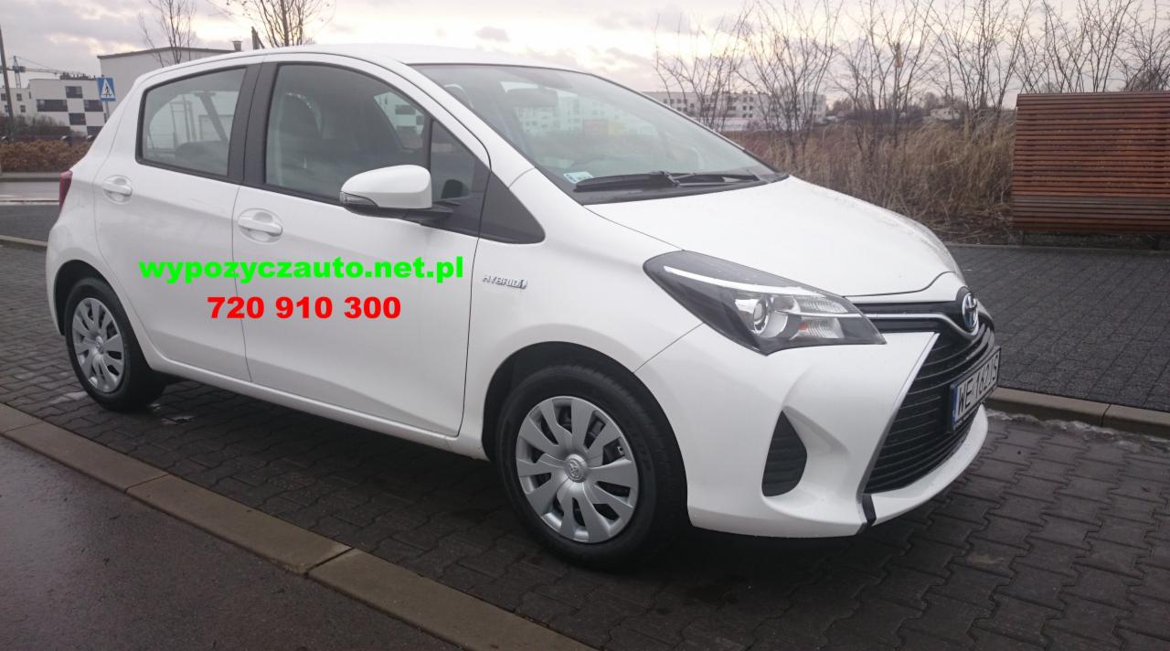 Biała Toyota Yaris Hybrid z numerem telefonu na boku, zaparkowana na ulicy. Widoczne budynki w tle i pochmurne niebo.