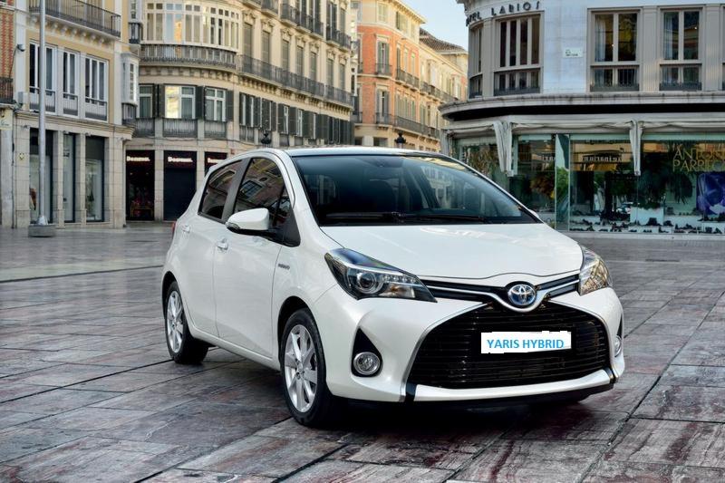 Biały Toyota Yaris Hybrid na tle miejskiej architektury z kamienną nawierzchnią. Auto zaparkowane na placu przed budynkami.