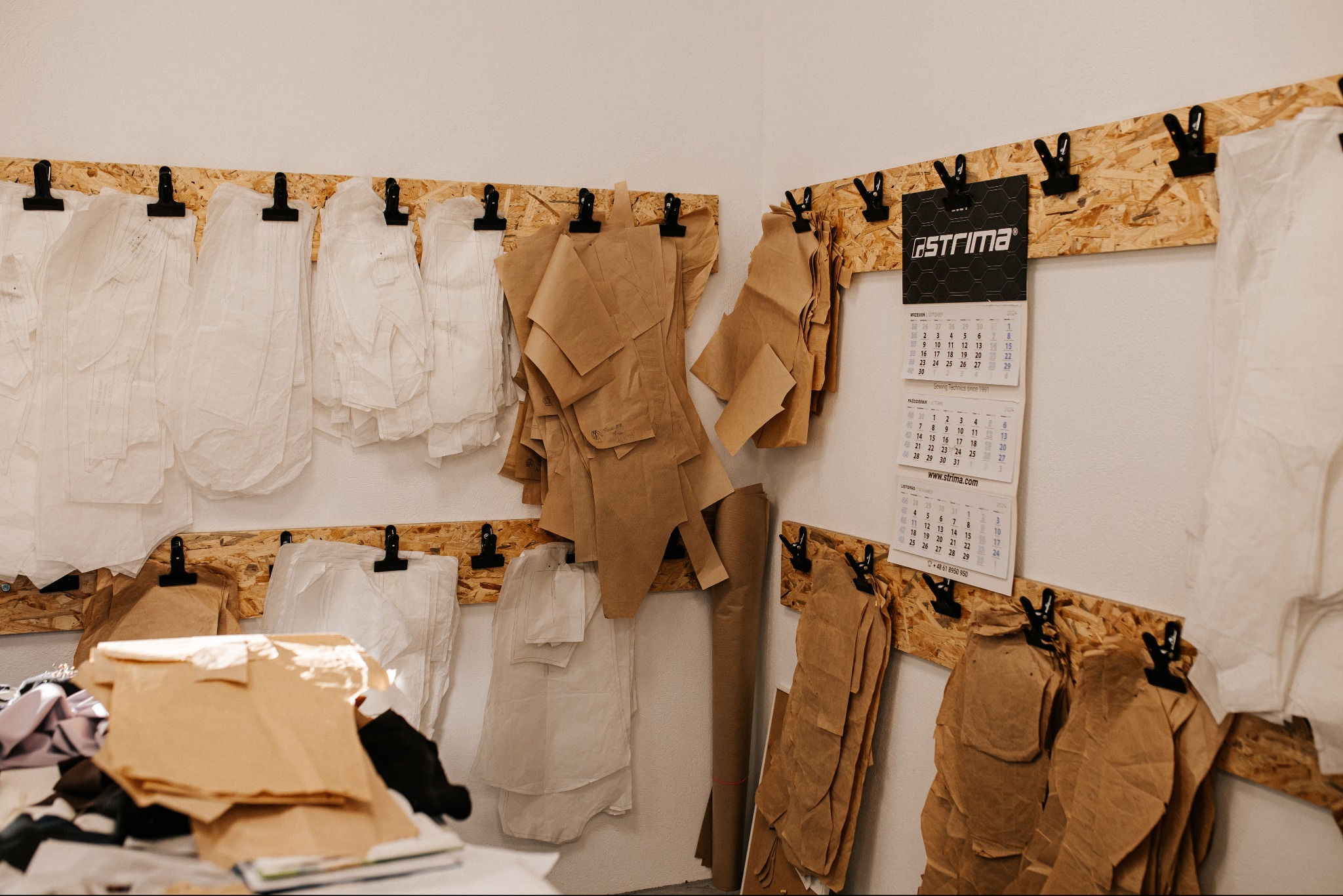 Pracownia krawiecka: Szablony krawieckie z papieru i transparentnej folii zawieszone na tablicach z płyty OSB; kalendarz firmowy Strima na ścianie.