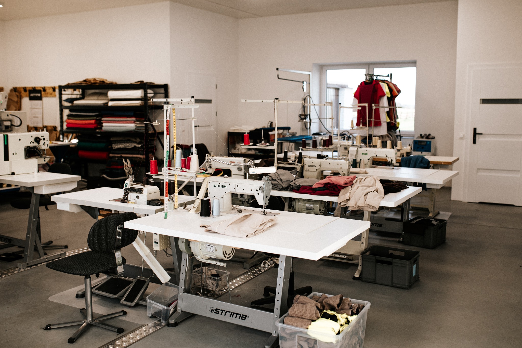 Jasne pomieszczenie z kilkoma stanowiskami pracy wyposażonymi w maszyny do szycia różnych marek (Siruba, Pfaff, Brother, Zoje), na stołach leżą fragmenty materiałów i nici, w tle regał z ułożonymi...