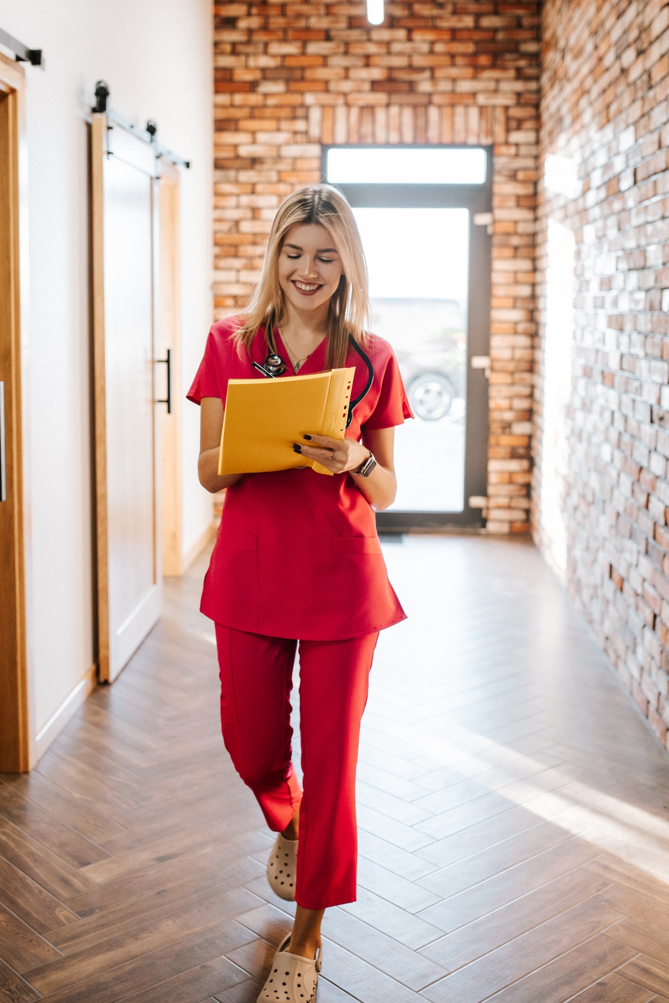 Kobieta w czerwonym uniformie, prawdopodobnie pracownik medyczny z Jutrosina, idzie korytarzem z żółtą teczką w rękach. Na szyi ma stetoskop, a w tle widać ceglaną ścianę.