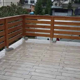 Wykończony balkon z drewnianą balustradą i płytkami imitującymi drewno. Widoczne zabezpieczenia na ścianie budynku. Tło stanowi zieleń i zabudowa.