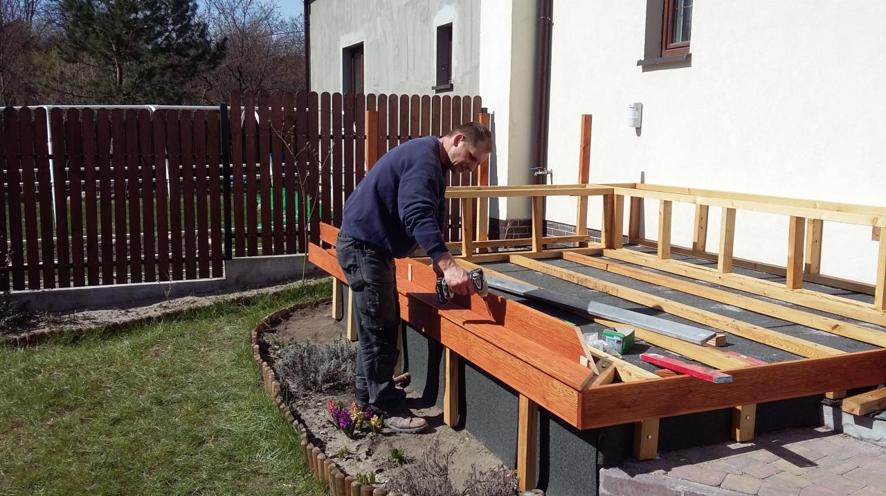 Budowa drewnianego tarasu przy domu: mężczyzna przykręca deskę w narożniku konstrukcji. Widoczne narzędzia, elementy konstrukcyjne i fragment ogrodu.
