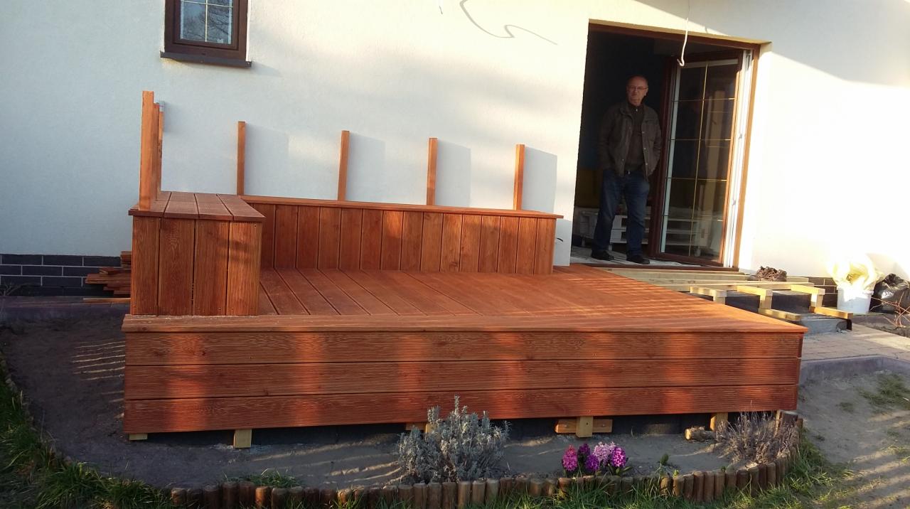 Drewniany taras w trakcie budowy, widoczne deski i elementy konstrukcyjne, mężczyzna w drzwiach. Świeże drewno, jasny budynek w tle, częściowo widoczne kwiaty.