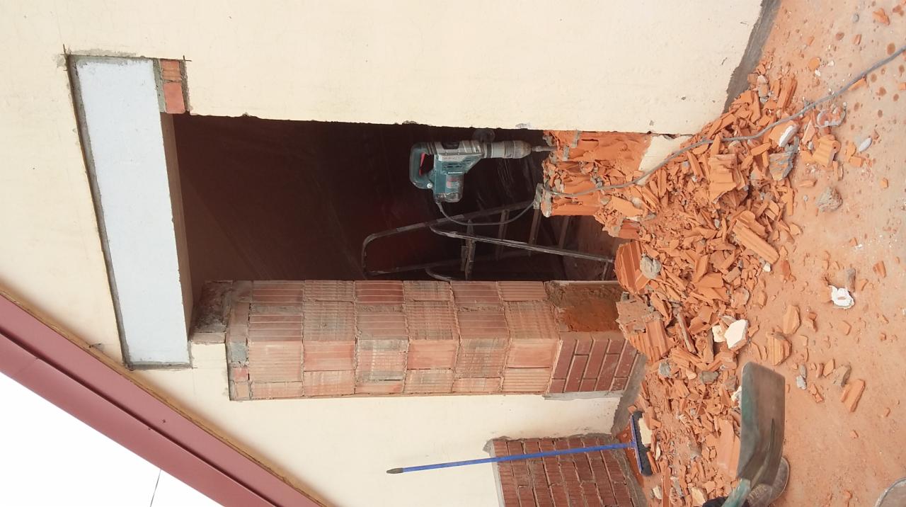 Demontaż fragmentu ściany z cegły przy użyciu młota udarowego. Widoczne gruz i narzędzia. Prace remontowe wewnątrz budynku z elementami konstrukcyjnymi dachu.