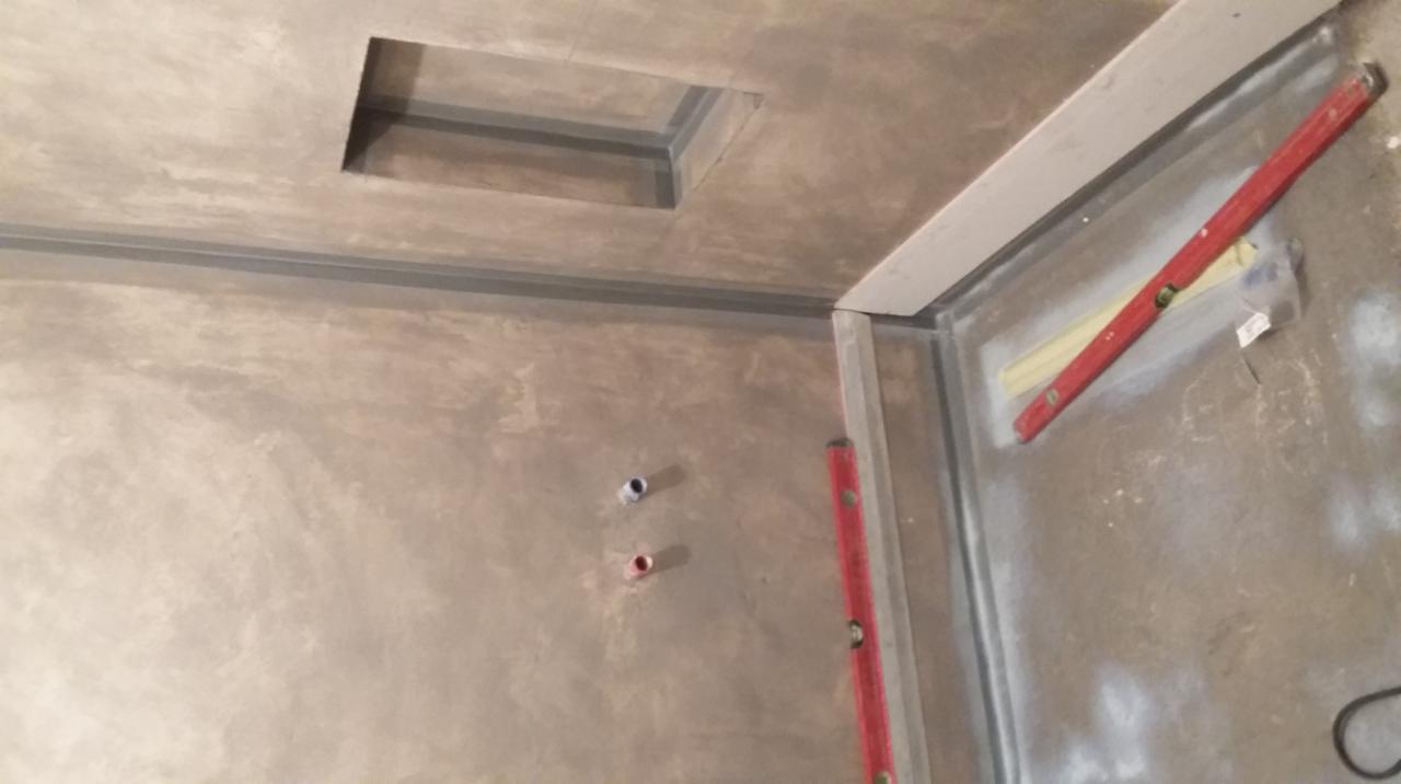 Surowa ściana z otworem w suficie, widoczne rury i poziomice. Przygotowania do wykończenia wnętrza, szare tynki, metalowe profile.