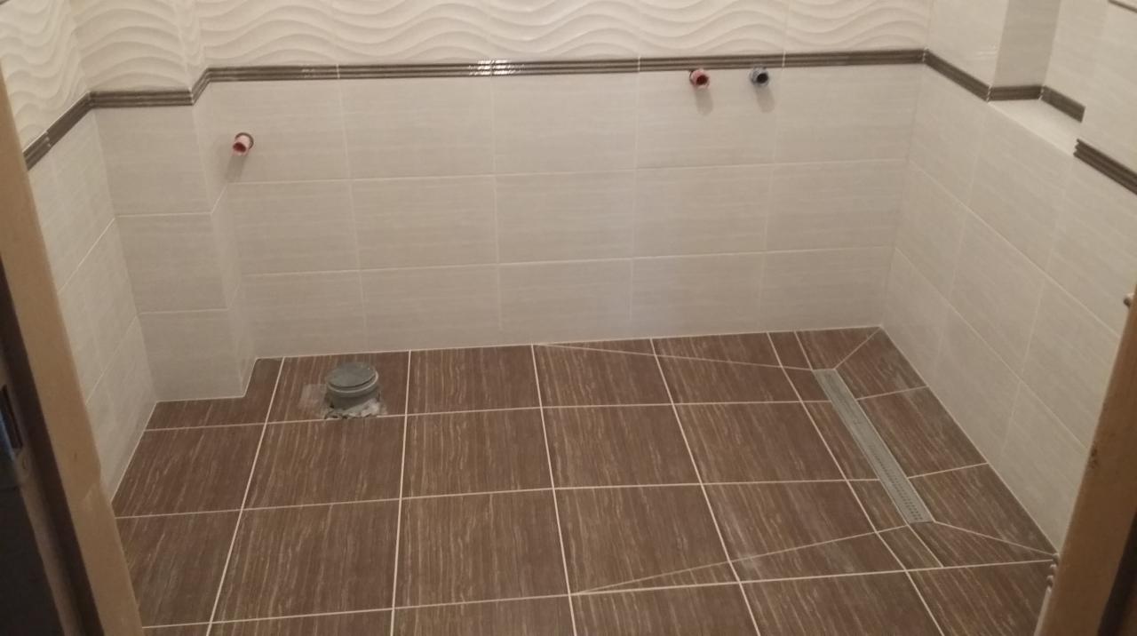 Wykończona kabina prysznicowa z brązowymi płytkami podłogowymi i beżowymi ściennymi z dekoracyjnym paskiem. Widoczny odpływ liniowy i instalacja wodna.