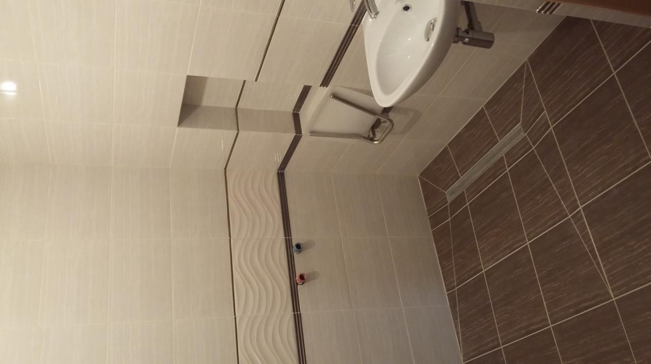 Wykończona łazienka z beżowymi i brązowymi płytkami, umywalką wiszącą i odpływem liniowym. Dekoracyjne, faliste płytki na ścianie. Widok na całą łazienkę.