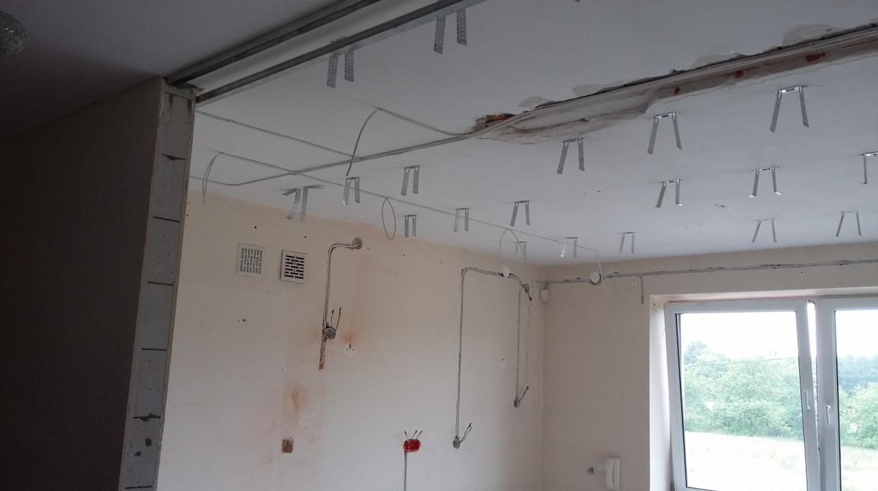 Montaż sufitu podwieszanego z widocznymi profilami, instalacją elektryczną i puszkami. Ściany w stanie surowym przed malowaniem. Okno z widokiem na zieleń.
