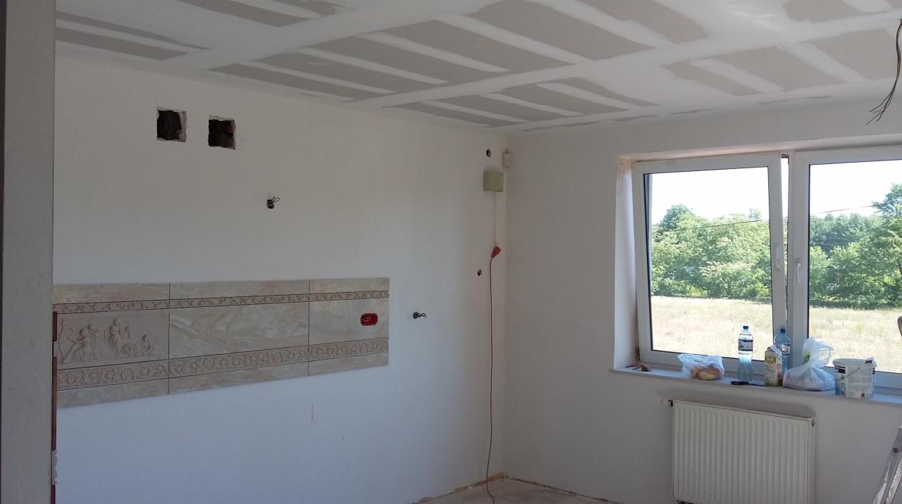 Wnętrze w trakcie prac: sufit z płyt gipsowych, fragment dekoracyjnych płytek na ścianie, okno z widokiem na zieleń. Widoczne przewody i instalacje.