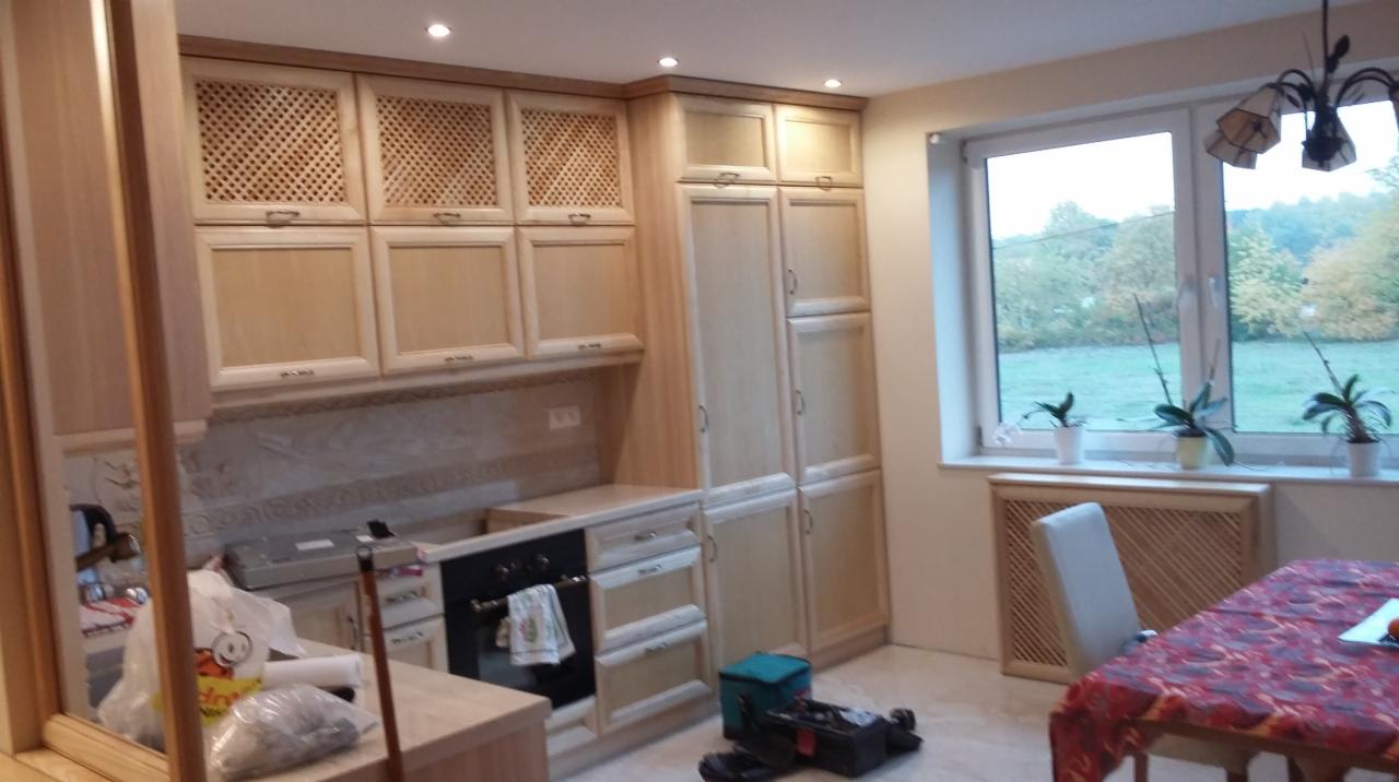 Jasna, drewniana zabudowa kuchenna z ażurowymi szafkami górnymi. Widoczne narzędzia i akcesoria, sugerujące etap montażu. Okno z widokiem na zieleń.