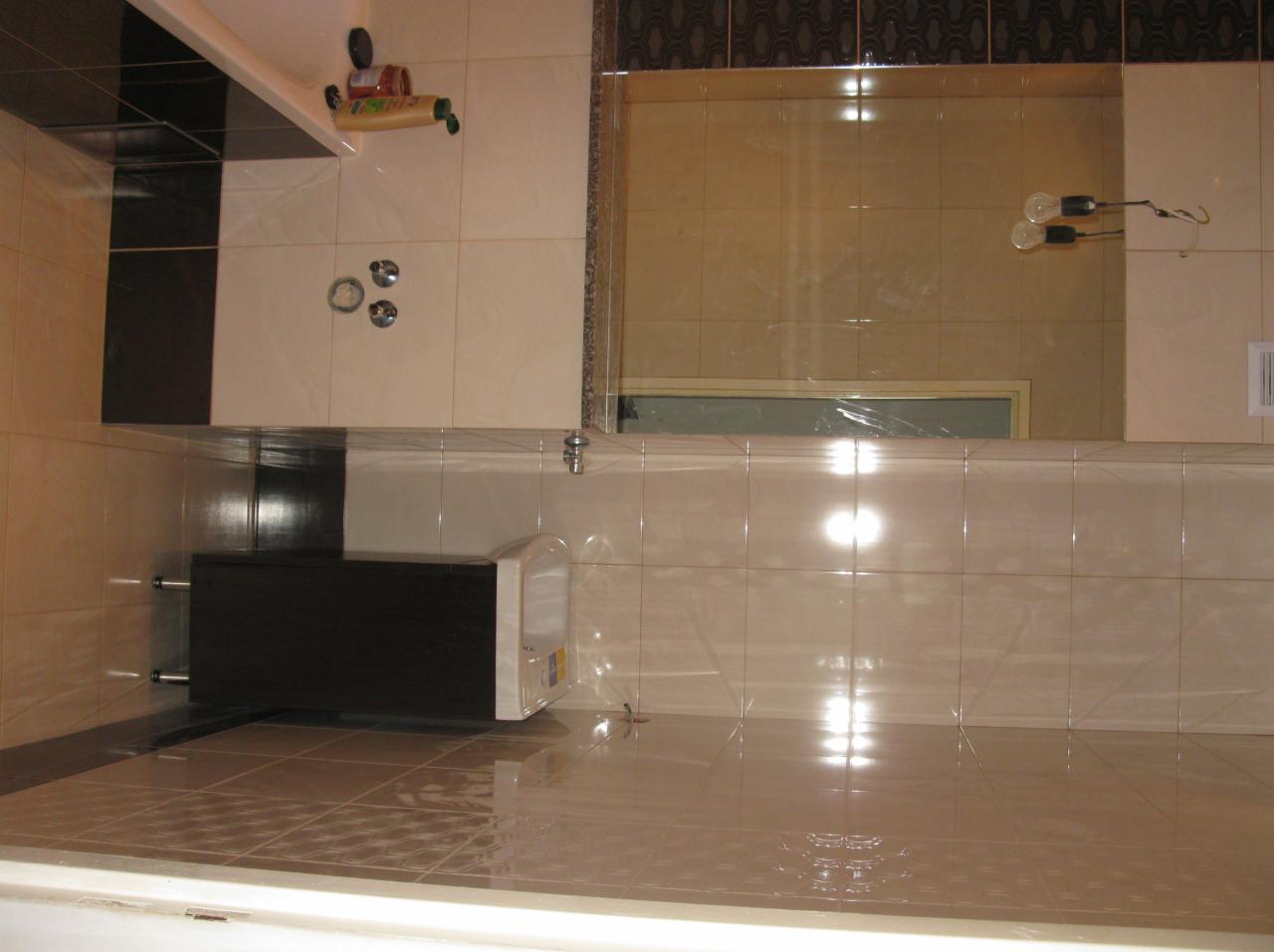 Wykończona łazienka z beżowymi płytkami, czarnym akcentem i lustrem. Widoczne akcesoria łazienkowe i fragment kabiny prysznicowej. Estetyczne i funkcjonalne wnętrze.