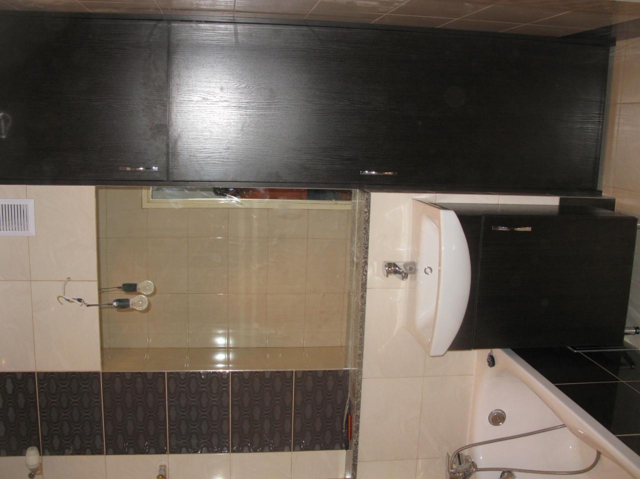 Wykończona łazienka z ciemnymi szafkami nad umywalką i wanną, kremowe płytki z dekoracyjnym pasem. Widoczne oświetlenie i wentylacja.