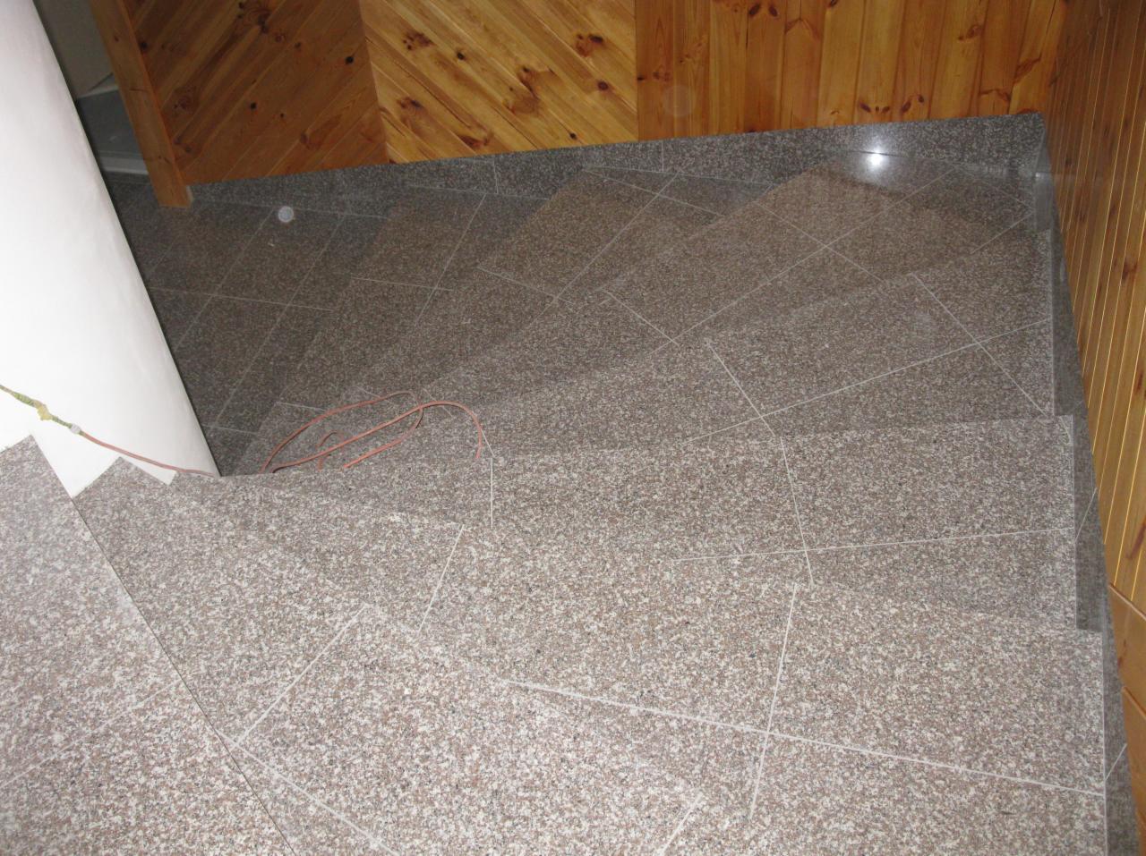 Wykończone schody z płytek o szarej, nakrapianej fakturze, prowadzące w dół. Widoczne drewniane panele na ścianie i wystający przewód elektryczny.