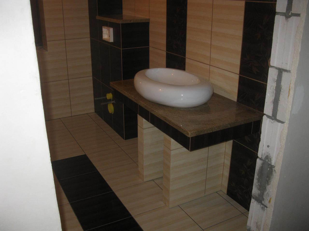 Nowoczesna łazienka z umywalką na blacie z kamienia i czarno-beżowymi płytkami. Widoczny fragment ściany w stanie surowym. Nietypowe wykończenie pod umywalką.