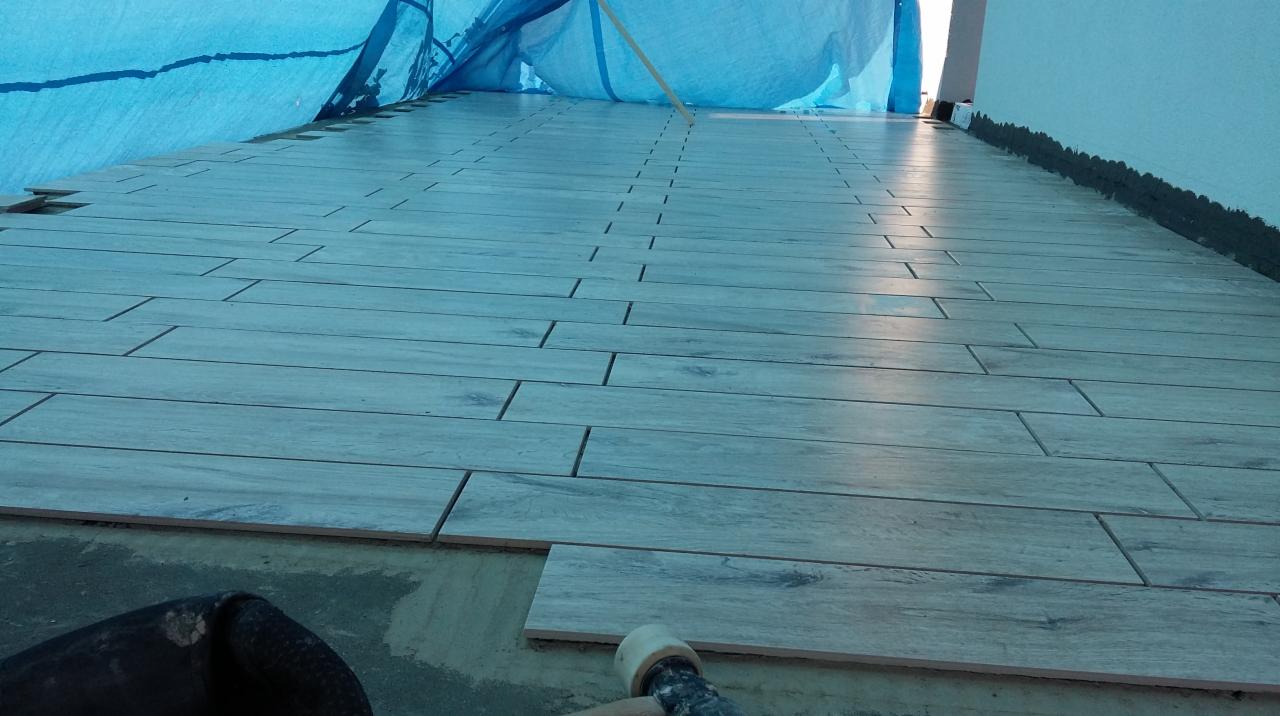 Układanie podłogi z płytek imitujących drewno, widoczne spoiny i narzędzia. Część podłogi przykryta folią ochronną, etap prac wykończeniowych.