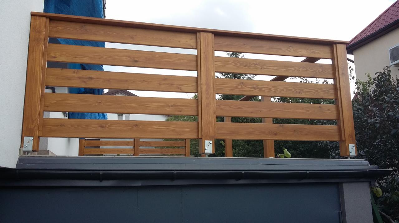 Drewniana balustrada balkonowa z poziomymi deskami, mocowana do ściany budynku. Widoczne metalowe elementy mocujące i fragment dachu garażu.