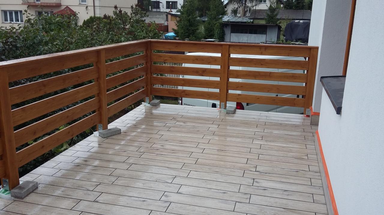 Wykończony balkon z drewnianą balustradą i płytkami imitującymi drewno. Widoczne zabezpieczenia na ścianie budynku. Tło stanowi zieleń i zabudowa.