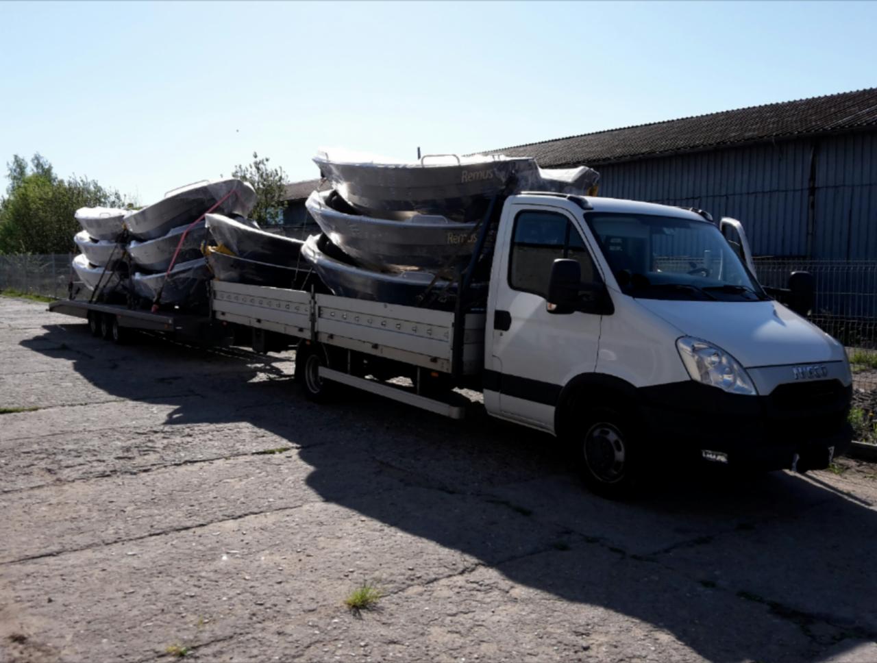 Biała ciężarówka Iveco z przyczepą przewozi kilka łodzi Remus, zabezpieczonych folią, na betonowym placu pod błękitnym niebem. Widoczna metalowa hala w tle.