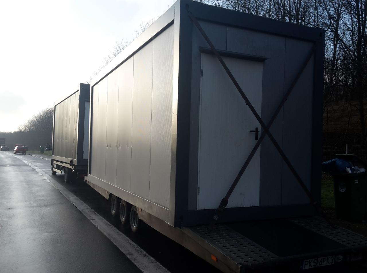 Specjalistyczny transport dwóch kontenerów modułowych na niskopodwoziowej przyczepie. Widoczna boczna ściana kontenera z drzwiami i zabezpieczeniami.