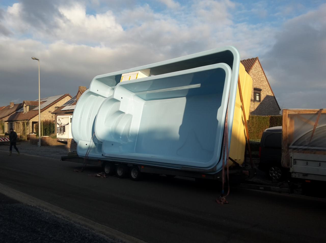 Transport basenów na przyczepie, widok z boku na tle zabudowy mieszkalnej. Jasnoniebieskie niecki basenowe zabezpieczone pasami transportowymi.