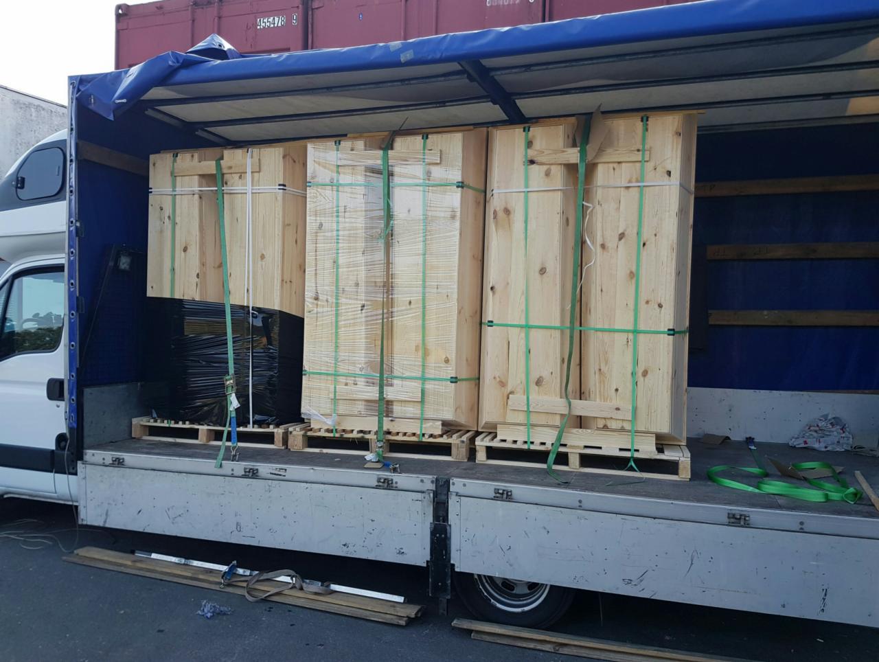 Załadunek trzech drewnianych skrzyń zabezpieczonych zielonymi pasami na paletach wewnątrz naczepy, widoczna biała kabina samochodu dostawczego i fragment czerwonego kontenera w tle.