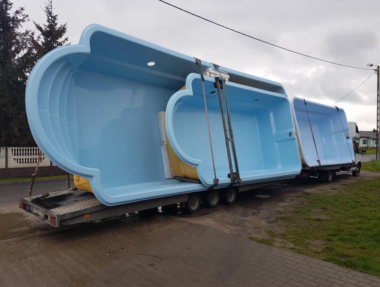 Trzy niebieskie, plastikowe baseny na przyczepach transportowych, zabezpieczone pasami, widoczne na zewnątrz w pochmurny dzień.